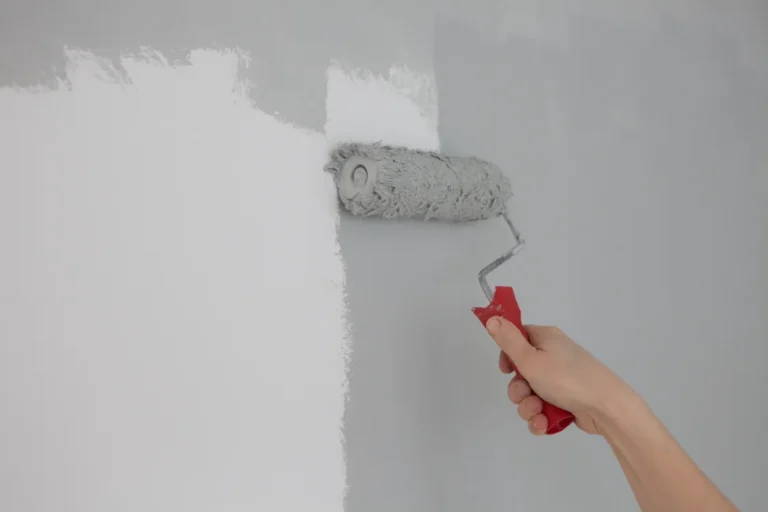 Interior painting project with a painter using a roller to apply light gray paint over a white wall.