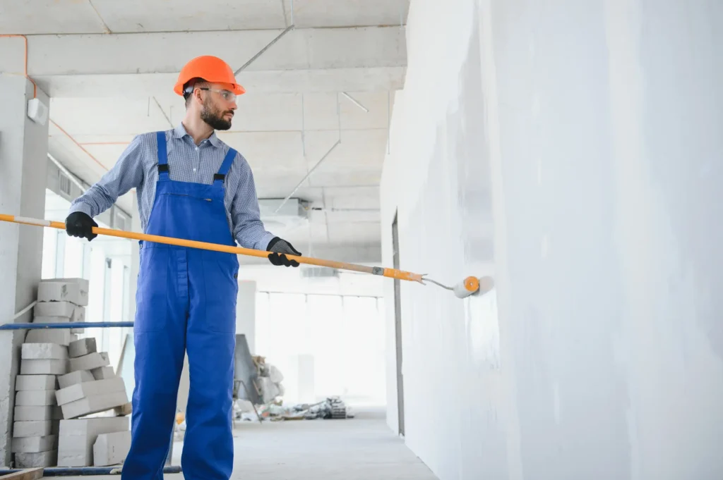 A man with overalls and a hat painting the primer on the walls.